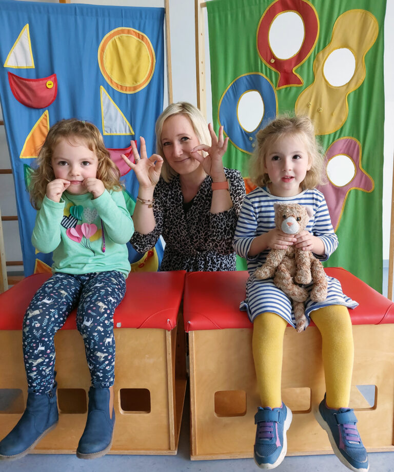 Amelie (links) und Erzieherin Yvonne Haverkamp „zeigen“ mit ihren Fingern Schnurrhaare, die Gebärde für „Katze“. Inga präsentiert das passende Stofftier dazu. Foto: Wagner